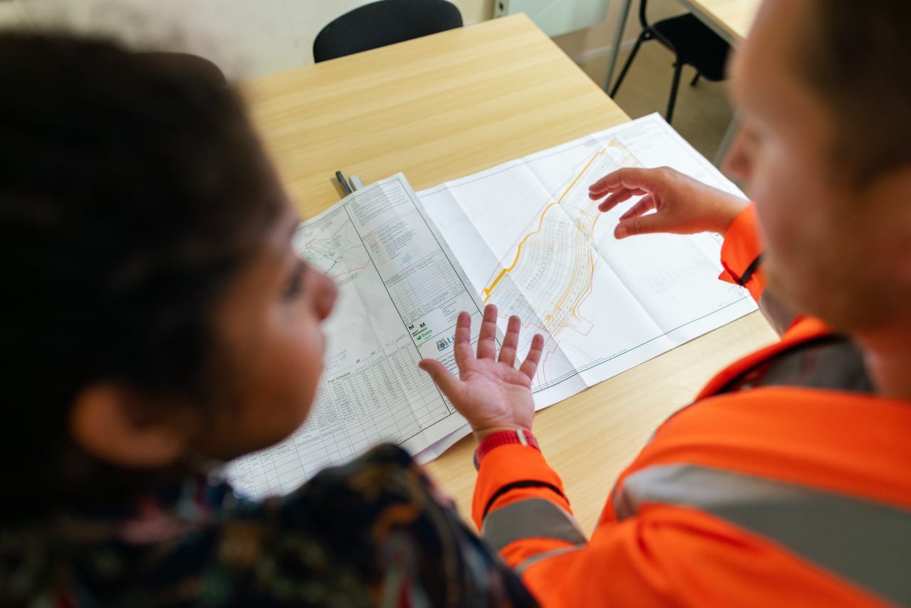 Two people, one in an orange high-visibility jacket, review a large technical drawing on an office table.