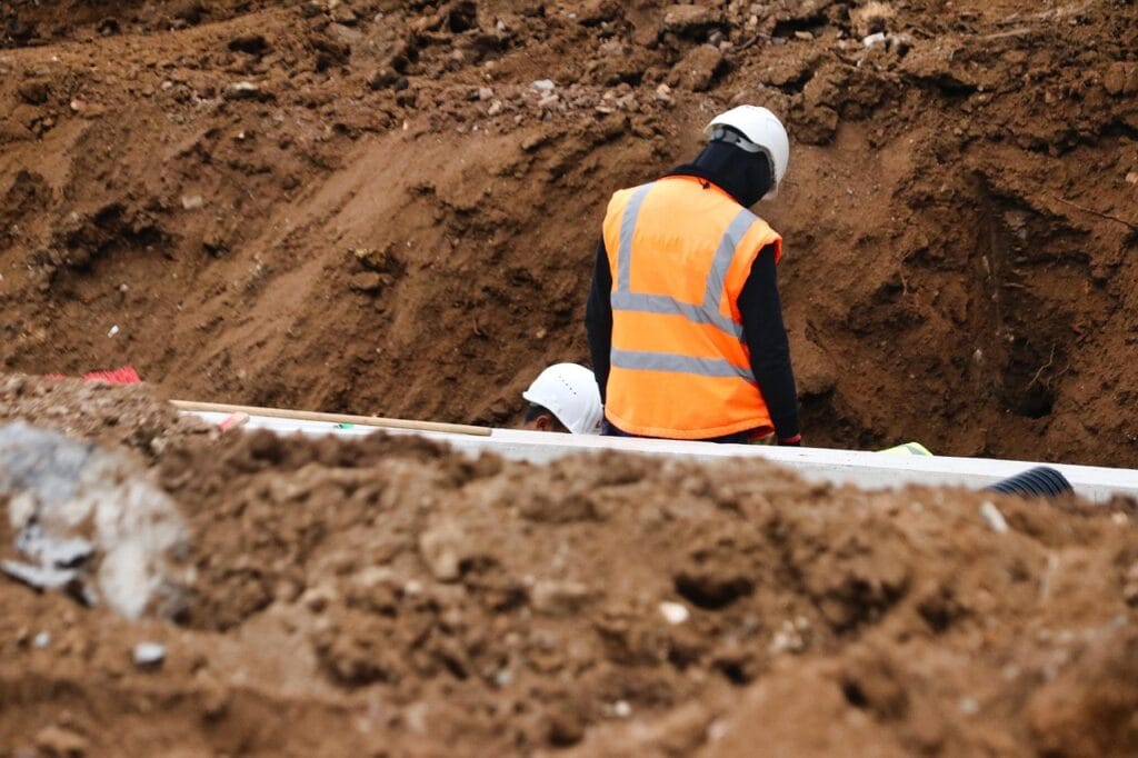 Workers in hard hats beside exposed utility pipes in an excavated trench, one wearing an orange reflective vest.