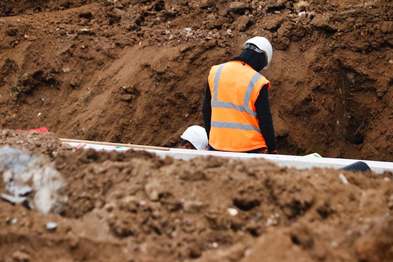 Workers in hard hats beside exposed utility pipes in an excavated trench, one wearing an orange reflective vest.